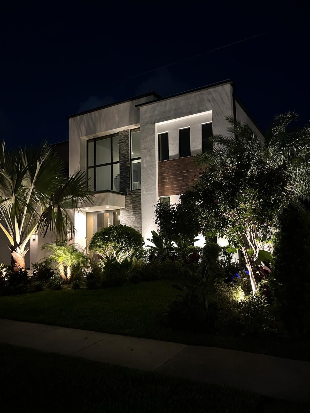 Modern Jupiter home with warm white landscape lighting highlighting palm trees and architectural features, built to withstand Florida's coastal climate.